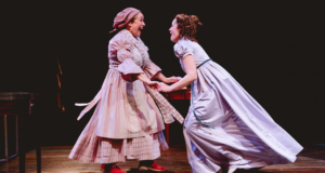 Gilly Tompkins and Paula Lane in long old fashioned dresses facing each other with excited looks on their faces