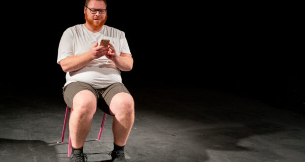 Plus-size white ginger man on stage, sitting on pink chair, looking at his mobile phone. Background is black.