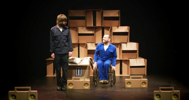 Daryl, a white man wearing a blue overall sits in his wheelchair. Next to him stands Jon, A white man wearing a dark blue overall. They are looking at each other. On the floor around them are radios made out of cardboard and behind them is a wall of boxes.