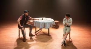 Two actors sit either side a round table on a bare stage, one leaning across the table talking to the other who sits with arms folded